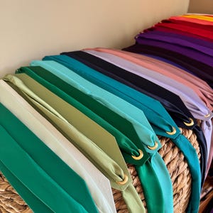 May include: A collection of long, smooth fabric strips in a variety of colors including white, green, teal, black, purple, red, and yellow. Each strip features a pointed end secured with a circular gold-toned metal grommet or eyelet.