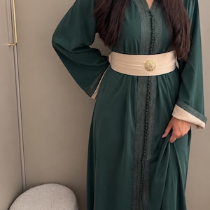 Puede incluir: Un caftán verde oscuro largo hasta el suelo con mangas largas y puños beige, escote en V y bordados intrincados en verde oscuro a lo largo de la parte frontal. Se lleva un cinturón ancho de color beige con una hebilla decorativa dorada.