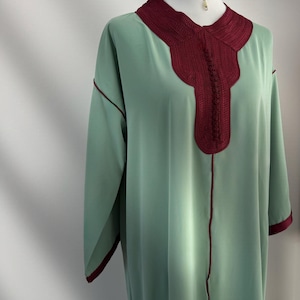 Op de afbeelding: Een mintgroene kaftan-stijl jurk met een bordeauxrode halslijn en bies, tentoongesteld op een mannequin. De jurk heeft lange mouwen en een centrale verticale naad. Een turquoise kralen choker ketting met een gouden hanger wordt gedragen.