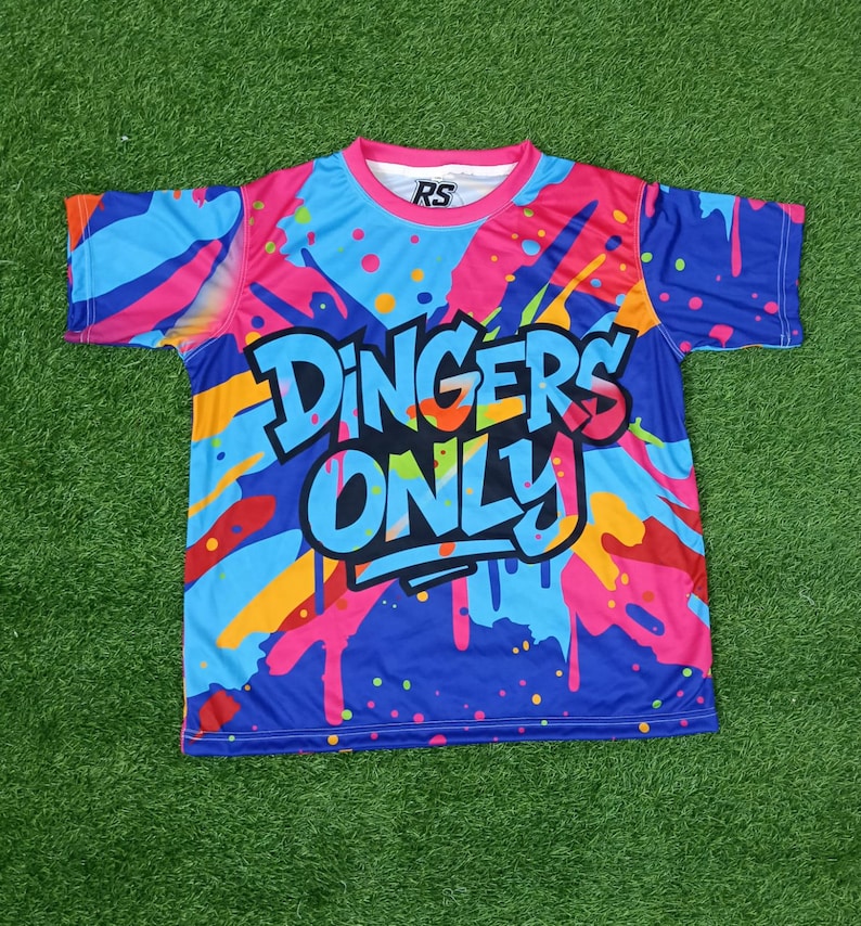 May include: A brightly coloured t-shirt with a blue base and a design of pink, orange, and yellow paint splashes. The text "DINGERS ONLY" is displayed in a bold, stylized font across the front of the shirt.