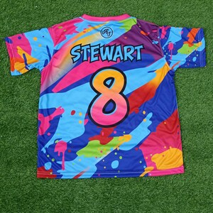 May include: A brightly coloured short-sleeved jersey with an abstract design. The jersey displays the name "STEWART" above the number "8". The colour scheme includes blue, pink, orange, and purple, with paint splatter effects.