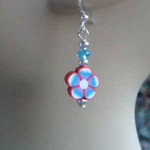 May include: A dangle earring featuring a red, white, and blue flower design. The flower is made of polymer clay and has a silver-colored metal hook. The earring also includes blue beads and silver-colored metal accents.