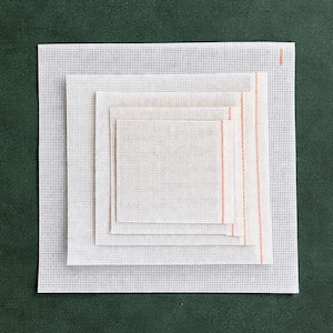 May include: A stack of five square needlepoint canvases in varying sizes. The canvases are white with a grid pattern, and the largest canvas is on the bottom. A vertical orange stripe is visible on the right side of the smallest canvas.