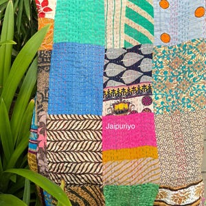 May include: A colourful patchwork textile, composed of various fabric squares stitched together. The design features a mix of blue, green, pink, orange, and brown hues. The textile is displayed outdoors, set against a backdrop of green foliage.