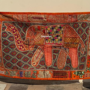 May include: A rectangular, orange textile wall hanging featuring an elephant design. The elephant is composed of various patterned fabrics in shades of orange, red, and gold. The border is intricately detailed with floral and geometric patterns.