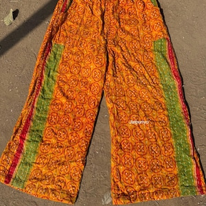May include: Wide-leg trousers in a vibrant orange hue, adorned with a repeating red circular design. Green and red stripes run down the sides. The fabric has a quilted appearance. The trousers are likely handmade.