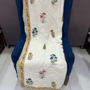 Peut inclure: Une couverture matelassée blanche avec des broderies florales orange, bleues et roses. La couverture est drapée sur une chaise bleue. La bordure présente un motif décoratif avec des accents jaunes et verts.