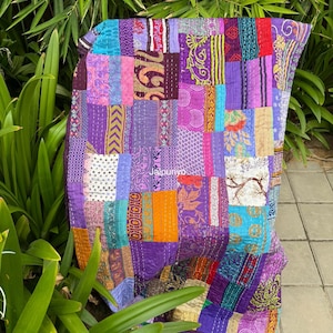 May include: A patchwork quilt in shades of purple, teal, orange, and pink. The quilt is made of various fabric pieces with different patterns and textures. The quilt is draped over a surface with green foliage in the background.