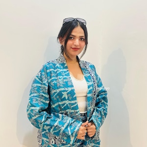 May include: A blue quilted jacket featuring a repeating floral pattern in shades of teal and white, with a contrasting dark blue and white geometric border along the front opening, worn over a white ribbed tank top and a blue denim mini skirt.