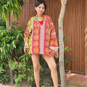 May include: A long-sleeved hooded pullover featuring a vibrant patchwork design with shades of pink, yellow, and green. The garment displays intricate floral and geometric patterns throughout, with a V-neckline and a loose, relaxed fit.