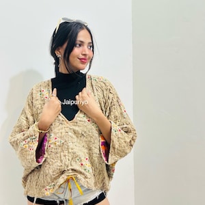 Pode incluir: Uma jaqueta acolchoada estilo quimono em bege com superfície texturizada, pequenos bordados geométricos coloridos, mangas largas e uma bainha com cordão e laços amarelos. O texto Jaipuriyo está impresso na parte frontal da peça.