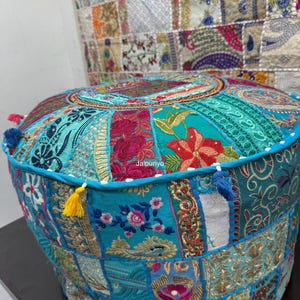 Puede incluir: Un puf redondo de patchwork en tonos turquesa, rosa y dorado. El puf presenta una variedad de parches de tela bordada con motivos florales y geométricos. Tiene un ribete azul y está adornado con pequeñas borlas de varios colores.