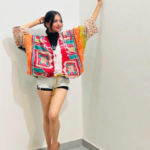 May include: A loose-fitting, open-front kimono jacket featuring a vibrant patchwork of orange, red, green, and white floral patterns. The garment has wide three-quarter length sleeves, a drawstring waist tie, and a visible Jaipuri brand label.