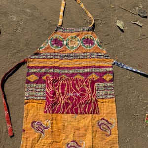 Könnte beinhalten: Bunter Patchwork-Schürze. Die Schürze hat eine Mischung aus orangefarbenen, gelben und lila Stoffen mit Paisley- und Blumenmustern. Mit Tasche und langen Bändern zum Binden.