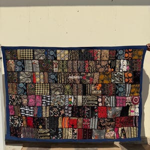 May include: A patchwork textile with a dark blue border. The textile is composed of numerous rectangular patches in various colors and patterns, including floral, geometric, and abstract designs. The word "Jaipuriyo" is visible on the textile.