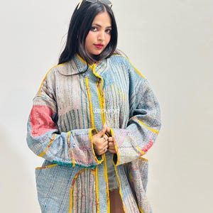 May include: A long-sleeved quilted jacket featuring a patchwork design with various shades of blue, white, and red accents. The garment has a bright yellow trim along the edges, a collar, and two large front pockets. The text Jajpuriyo is visible.