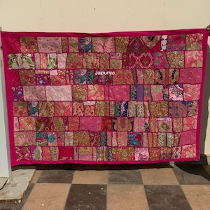 Peut inclure: Textile rectangulaire en patchwork dans les tons de rose, fuchsia et or. Le textile est composé de nombreux carrés de tissu avec divers motifs et textures. Les bords sont encadrés d'une bordure fuchsia unie. Le texte "Jaipuriyo" est visible.