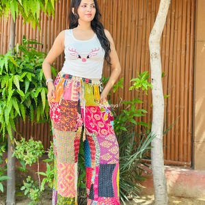 May include: A white sleeveless tank top featuring a pink reindeer graphic and wide-leg trousers made from a patchwork of multicoloured fabrics with various intricate patterns, including floral, geometric, and paisley designs in shades of red, yellow, and blue.