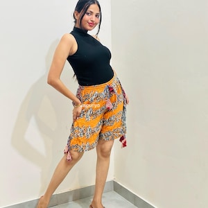 May include: A black sleeveless mock neck top paired with mustard yellow shorts featuring a blue and white floral pattern, an elastic waistband with a drawstring, and decorative tassels on the sides, worn with brown woven wedge sandals.
