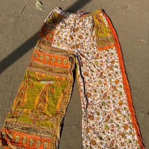 Può includere: Pantaloni a gamba larga con design patchwork. I pantaloni presentano un mix di motivi, tra cui paisley e floreali, nelle tonalità arancione, verde e bianco. Il testo "Jaipuriyo" è visibile.