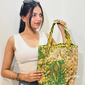 May include: A tote bag featuring a cream-colored base with a green and yellow floral paisley pattern. The bag includes two fabric handles and a square front pocket with visible stitching. The word Jaipuriyo is printed in the bottom right corner.