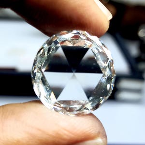 Puede incluir: Una gema clara y facetada con un dise&ntilde;o de talla rosa. El diamante de forma redonda se sostiene entre los dedos, mostrando sus intrincadas facetas y patrones triangulares internos. La gema refleja la luz, resaltando su claridad y brillo.