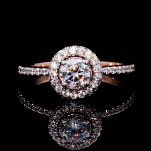 Puede incluir: Un anillo de compromiso de oro rosa con una piedra central grande, redonda y transparente rodeada de piedras más pequeñas. La banda del anillo también está adornada con pequeñas piedras, sobre un fondo negro, con su reflejo visible.