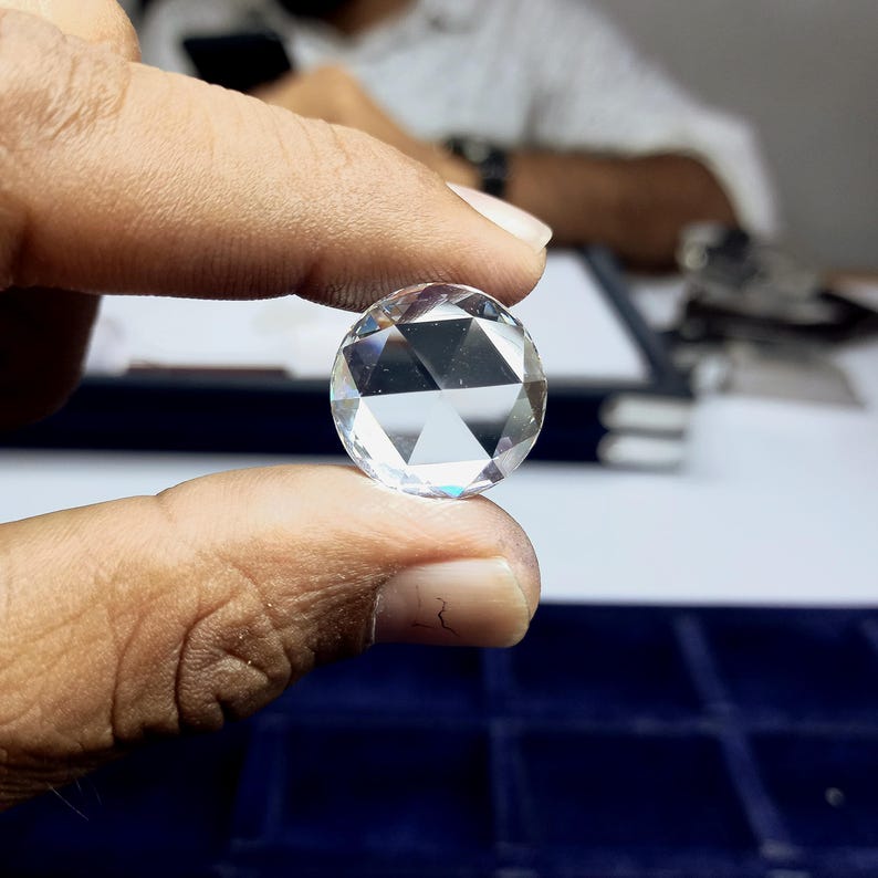 Puede incluir: Una gema clara, de talla redonda, con m&uacute;ltiples facetas, sostenida entre los dedos. La gema brilla, reflejando la luz. Es un diamante, una piedra preciosa que se usa a menudo en joyer&iacute;a.
