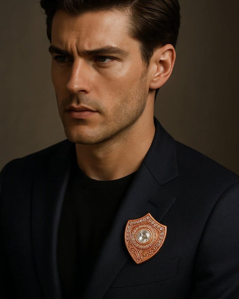 Puede incluir: Broche en forma de escudo de color cobre con una gema redonda y transparente en el centro. La broche est&aacute; adornada con gemas transparentes m&aacute;s peque&ntilde;as alrededor de la piedra central y a lo largo de los bordes. Se sujeta a una blazer azul marino oscuro.