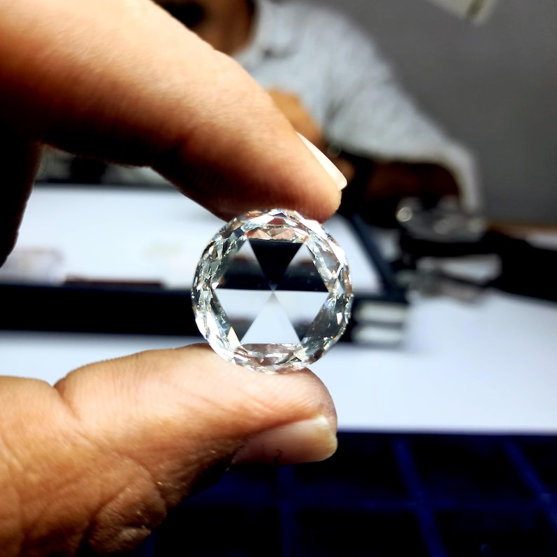 Puede incluir: Una gema clara y facetada con un dise&ntilde;o de talla rosa. El diamante de forma redonda se sostiene entre los dedos, mostrando sus intrincadas facetas y patrones triangulares internos. La gema refleja la luz, resaltando su claridad y brillo.