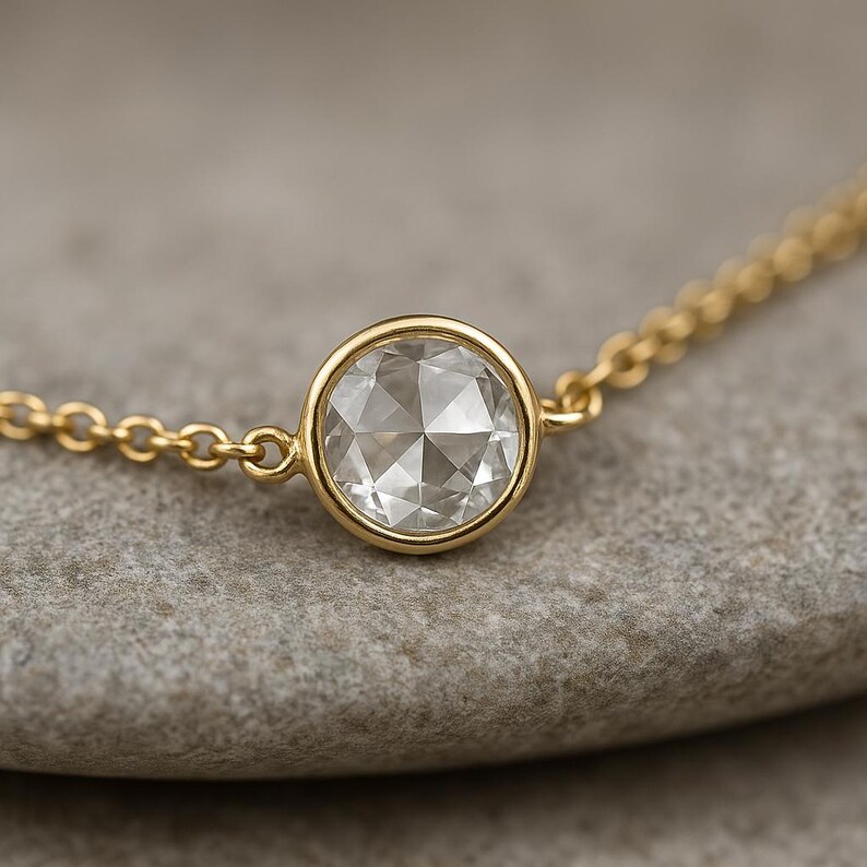 Puede incluir: Una delicada pulsera de cadena de oro con un diamante redondo y facetado engastado en un bisel de oro. El diamante brilla con m&uacute;ltiples facetas, reflejando la luz. La pulsera se muestra sobre una superficie gris neutra.