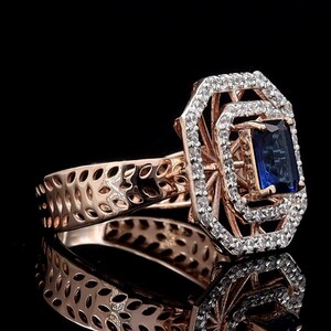 Puede incluir: Anillo de oro rosa con una piedra preciosa rectangular azul oscuro, rodeada de pequeñas piedras preciosas claras. La banda del anillo tiene un diseño calado. El anillo está sobre un fondo negro, con su reflejo visible.
