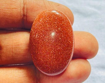 May include: An oval-shaped cabochon of goldstone, featuring a deep reddish-brown base color densely packed with tiny, reflective metallic copper-colored flecks that create a shimmering, glittery appearance across the smooth, polished surface.