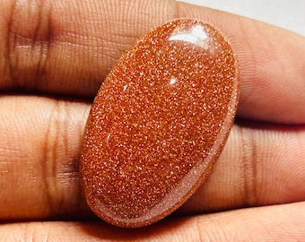 May include: Oval-shaped, polished gemstone with a sparkling, golden-brown appearance. The stone has a smooth, glossy surface and is held in a hand. The gemstone's color is a warm, shimmering hue, with tiny reflective particles.