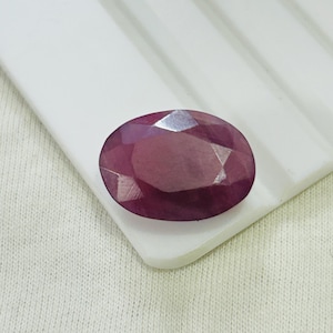 May include: A faceted, oval-shaped ruby gemstone. The gem is a deep, rich red colour with multiple facets that reflect light. The ruby is set against a white surface with a textured pattern.