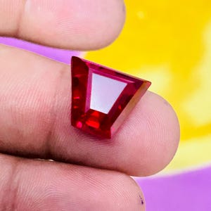 May include: A faceted, trapezoidal-shaped gemstone with a deep, vibrant red color. The stone features a polished surface with sharp, geometric edges and a clear, transparent quality that reflects light across its multiple angled facets.