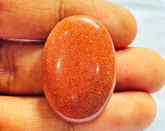 May include: An oval-shaped goldstone cabochon featuring a deep reddish-orange hue with a dense, sparkling metallic glitter effect throughout the smooth, polished surface of the gemstone.