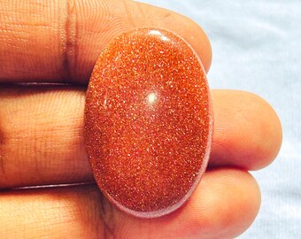 May include: An oval-shaped goldstone cabochon featuring a deep reddish-orange hue with numerous embedded metallic copper-colored flecks that create a reflective, shimmering appearance across the smooth, polished surface of the gemstone.