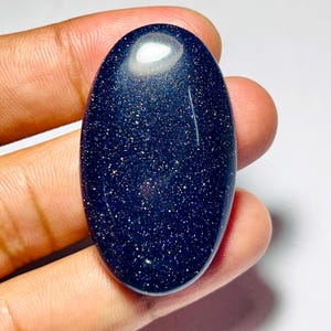 May include: An oval-shaped, polished blue sandstone gemstone with sparkling inclusions. The stone is held in a hand, showcasing its smooth, reflective surface and the tiny, glittery particles that give it a starry appearance. The gemstone is a deep, rich blue color.