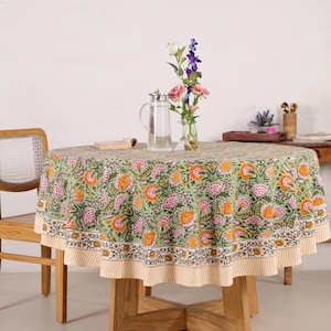 Puede incluir: Un mantel redondo con un estampado floral en tonos verdes, rosas y naranjas. El mantel está sobre una mesa de madera, adornado con un jarrón de flores y una jarra de agua. El mantel tiene un borde a rayas amarillas y blancas. La mesa mide unos 120 cm de diámetro.