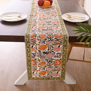 Puede incluir: Un camino de mesa floral con fondo blanco y un patrón repetido de flores naranjas y rosas. El camino de mesa tiene un borde verde con un diseño floral repetido. El camino de mesa está colocado sobre una mesa de madera marrón oscuro.