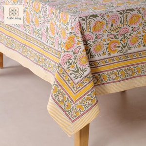 Puede incluir: Un mantel floral con fondo blanco y un patrón repetido de flores amarillas y rosas. El borde es amarillo con un diseño a rayas. El mantel está sobre una mesa de madera.