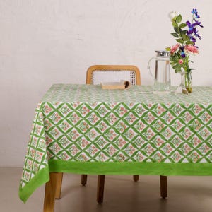 Puede incluir: Un mantel con estampado floral verde y blanco con un borde verde brillante. El mantel está sobre una mesa de madera. Una jarra de cristal transparente y un jarrón con flores están sobre la mesa.