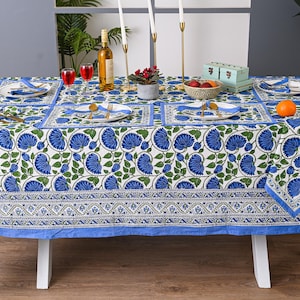 Puede incluir: Un mantel rectangular con un estampado floral azul, preparado para una comida. La mesa está puesta con platos, servilletas y cubiertos. Una botella de vino, velas y un cuenco de fruta también están sobre la mesa. La mesa mide 152 cm de ancho y 203 cm de largo.