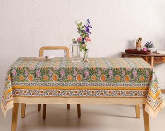 May include: A rectangular tablecloth with a floral pattern in green, yellow, and pink. The tablecloth is on a wooden table. A vase of flowers and a pitcher are on the table. The background is light-coloured.