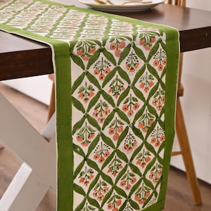 Può includere: Runner da tavolo floreale verde e bianco. Il runner presenta un motivo ripetuto di foglie verdi e fiori arancioni su sfondo bianco, con un bordo verde pieno. È esposto su un tavolo di legno scuro, con un piatto e posate.