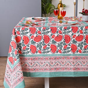 Puede incluir: Un mantel blanco con un estampado floral rojo y detalles verdes. La mesa está puesta con platos, cubiertos y copas de vino. También hay una botella de vino y velas. El mantel es de tela estampada.