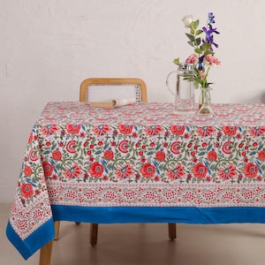 Puede incluir: Un mantel rectangular blanco con un vibrante estampado floral en rojo, verde y azul. El mantel tiene un borde azul y cubre una mesa de madera. Un jarrón con flores y una jarra de agua están sobre la mesa.
