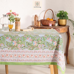 Puede incluir: Un mantel verde floral con un borde detallado, colocado sobre una mesa de madera. La mesa está adornada con una jarra, flores y una cesta de naranjas. El mantel presenta un patrón repetido de flores en tonos azules, naranjas y blancos.