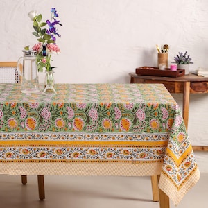 May include: A rectangular tablecloth with a floral pattern in shades of green, pink, and yellow. The tablecloth is draped over a wooden table. A yellow and white striped border is visible. A vase of flowers and a pitcher of water are on the table.